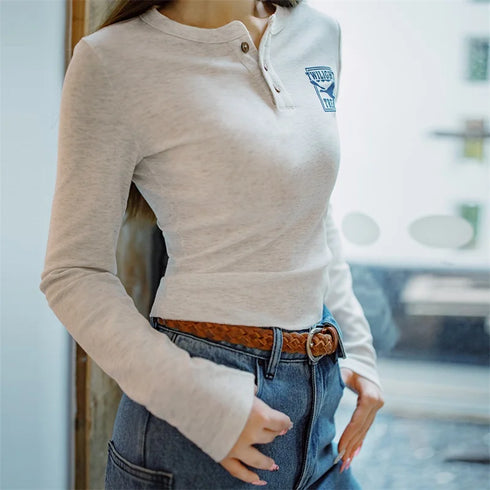 Person wearing a beige long-sleeve shirt with a logo and blue jeans, standing indoors.